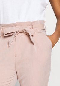 Ljust rosa byxor med ett bälte som har en rosett, veckad midja och sidofickor; tillverkade av ett texturerat tyg med ett subtilt randmönster.