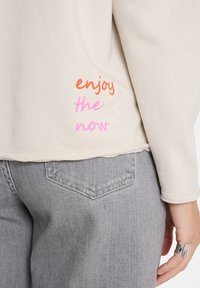 Beige Sweatshirt mit aufgesticktem "Enjoy the Now" in Orange und Pink, kombiniert mit grauen Jeans und einer Hand, die zwei silberne Ringe trägt.