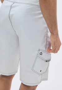 Fehér fürdőnadrág fekete varrással, oldalzsebbel és felhajtós fedéllel, valamint a zseb felett található logófolttal. Simább, könnyű anyagból.