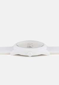 Montre blanche avec un boîtier rond, un cadran texturé et un verre transparent. Elle dispose d'un bracelet en silicone blanc et d'une couronne de remontoir proéminente.