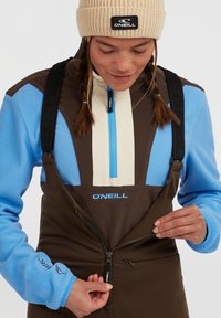 Maglione in pile con colori blu, marrone e crema; salopette marroni con zip; logo O'Neill visibile; modello che chiude la zip delle salopette.
