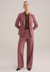 Rosa maßgeschneiderte Blazer, kombiniert mit passenden hoch taillierten Hosen, getragen über einem braunen Poloshirt, abgerundet mit spitz zulaufenden Schuhen.