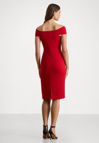 Κόκκινο φόρεμα με ανοιχτούς ώμους, εφαρμοστό, με φερμουάρ στην πλάτη, μήκους μέχρι το γόνατο με σκίσιμο, φορεμένο με μαύρες ψηλές γόβες.