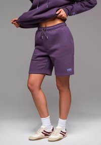Lila Baumwollshorts mit einem elastischen Bund und Kordelzug. Mit Taschen und einem kleinen Logopatch an der Seite. Kombiniert mit weißen Socken und Turnschuhen.