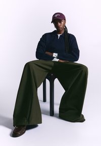 Oversized olive green pants, navy sweater, white shirt under, and a maroon cap with text. Brown shoes complete the look. Minimalist background.