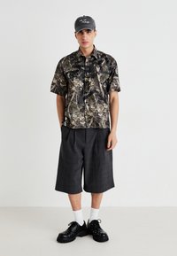 Cămașă scurtă cu imprimeu camuflaj și accente negre, asortată cu pantaloni scurți plisati gri închis. Pantofi negri cu ținte și șosete albe până la gleznă completează ținuta.