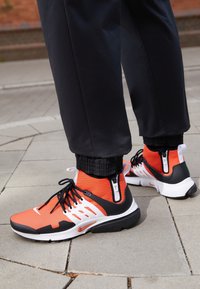 Osoba v černých kalhotách a jasně oranžových, černých a bílých sportovních teniskách se zipy vzadu, stojící na dlažbě venku.