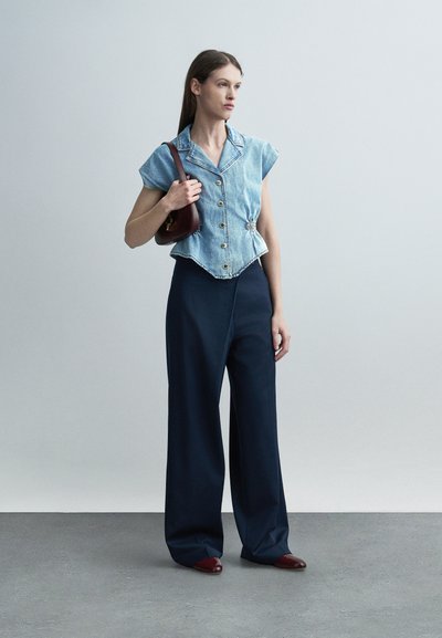 Femme portant un chemisier sans manches en jean, un pantalon large bleu marine, des chaussures bordeaux, tenant un sac à bandoulière bordeaux assorti, debout sur un sol gris.