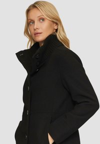 Schwarzer Mantel mit geripptem Kragen, großen Knöpfen und seitlichen Taschen. Der Stoff hat eine strukturierte Optik.