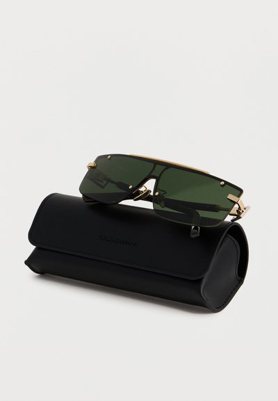 Ορθογώνια γυαλιά ηλίου με πράσινους φακούς και χρυσό πλαίσιο, τοποθετημένα πάνω σε μαύρη δερμάτινη θήκη Dolce & Gabbana σε λευκό φόντο.