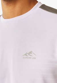 Fehér sportpóló, amely texturált hálós anyagból készült, kerek nyakkivágással és szürke díszítéssel a vállakon. A logó felirata: "CLOSURE LDN."