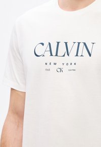 Camiseta blanca de algodón de Calvin Klein con un gran texto azul marino que dice "CALVIN NEW YORK 1968" y "CK", con un cuello redondo clásico.