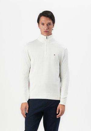 Een man in een witte lange mouwen pullover met een halve rits en een klein logo op de borst, gecombineerd met donkere broek.