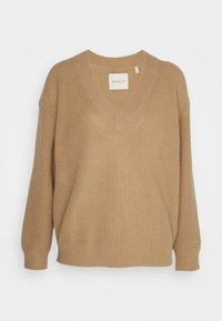 Pull beige tricoté avec un col en V, des épaules tombantes et des poignets côtelés, présentant un tissu texturé et une étiquette sur l'encolure.