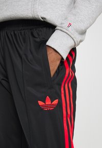 Pantalon de sport noir avec des bandes latérales rouges et un logo Adidas rouge brodé. Fabriqué en matériau lisse et léger ; comporte deux poches latérales.
