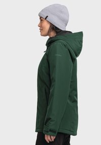 Grüne wasserdichte Jacke mit Kapuze, die eine glatte Textur, einen Reißverschluss und verstellbare Bündchen aufweist. Kombiniert mit einer hellgrauen Strickmütze.