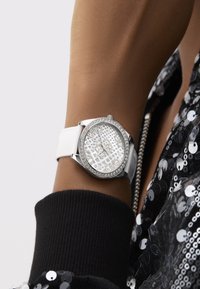 Valge nahkkummiga kell hõbedase korviga, sädelevate kristallaktsentidega ja tekstureeritud dialiga, millel on reljeefsed numbrid.