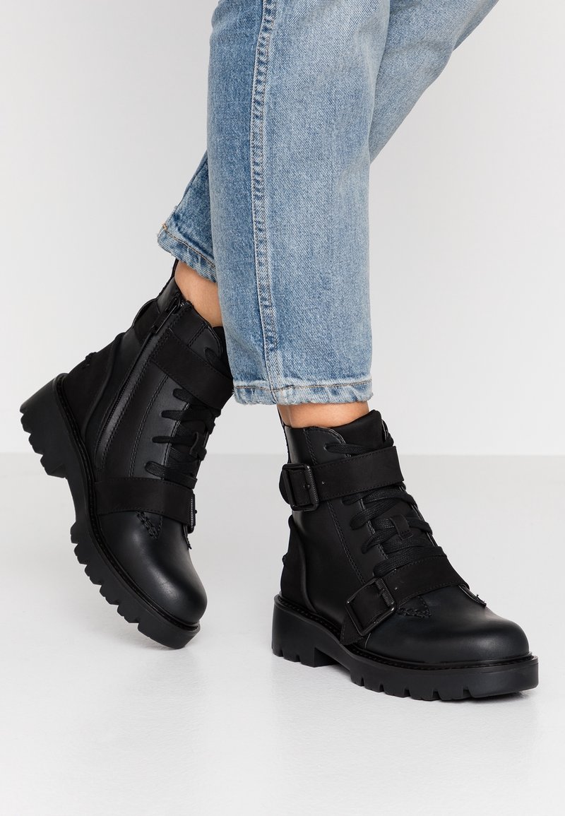 Czarne skórzane botki za kostkę z grubą podeszwą, wyposażone w sznurowadła, dwa paski z klamrami i teksturowaną cholewkę. W połączeniu z podwiniętymi jasnoniebieskimi dżinsami.