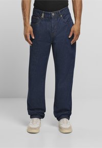 Mies, jolla on yllään väljät tummansiniset farkut ja valkoiset tennarit, seisoo kädet rentoina sivuilla yksiväristä taustaa vasten.