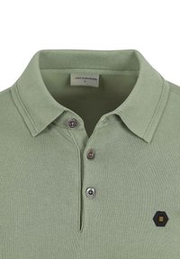 Groene polo gemaakt van ribbelstof, met een opgevouwen kraag, twee metalen knopen en een hexagonaal logo aan de linkerkant.