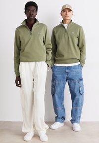 Twee personen dragen casual outfits: de één in een groene fleece met rits en witte broek; de ander in een groene fleece met rits en blauwe cargo jeans. Witte sneakers.