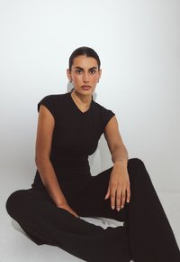 Moteris su tamsiais plaukais surištais atgal, vilkinti juodomis trumpomis rankovėmis ir plačiomis kelnėmis, sėdinti sukryžiuotomis kojomis prie baltos sienos.