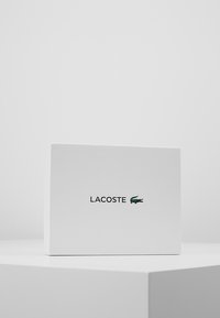 Biely textúrovaný box s čiernym logom Lacoste a malým zeleným krokodílom. Jednoduchý obdĺžnikový dizajn.