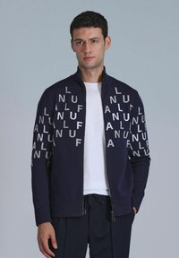 Marine blauwe zip-up jack met contrasterende zilveren letters op de mouwen, geribbelde manchetten en kraag. Gekenmerkt over een witte T-shirt, casual ontwerp.