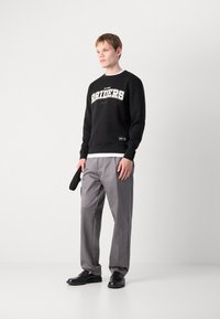 Sweatshirt de algodão preto com gráfico "RAIDERS" em branco, combinado com calças cinzas e sapatos pretos. Homem segurando um acessório preto.