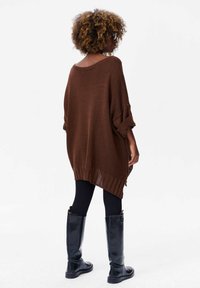 Bruine oversized gebreide trui met brede mouwen, ribgebreide onderrand en zijsplitsingen, gedragen over zwarte legging en kniehoge zwarte laarzen.