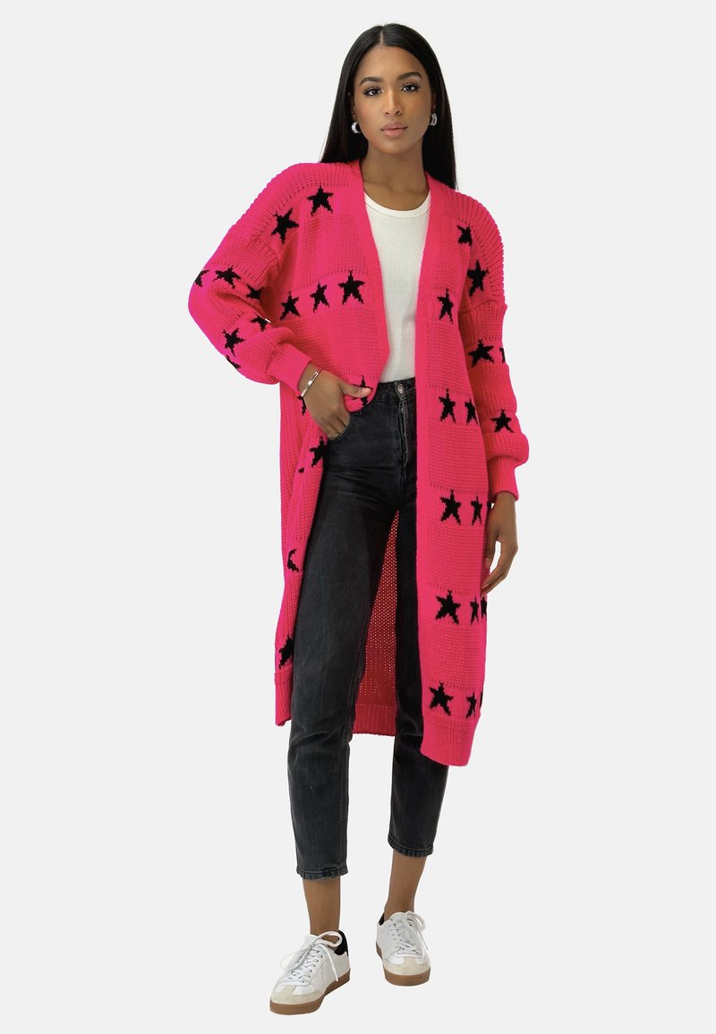 Long gilet en tricot rose à motifs d'étoiles noires, design à l'avant ouvert, poignets côtelés et coupe ample. Porté par-dessus un haut blanc et un jean foncé.