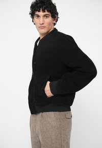 Νεαρός άνδρας με σγουρά σκούρα μαλλιά, φορώντας μια μαύρη ζακέτα bomber και μπεζ υφασμάτινο παντελόνι, ποζάροντας με τα χέρια στις τσέπες της ζακέτας.