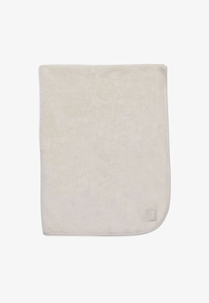 Zachte, beige pluche deken met een gladde textuur, afgeronde hoeken en een klein merklabel in de rechter benedenhoek.