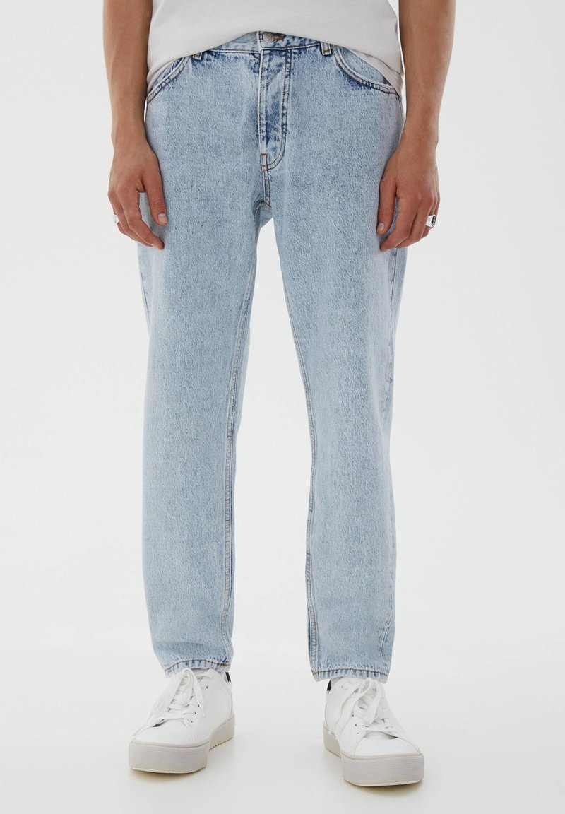 Personne portant un jean en denim bleu clair, des baskets blanches et une chemise blanche, debout devant un fond blanc uni.