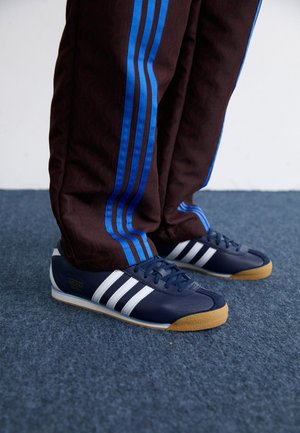 Osoba nosząca ciemne spodnie dresowe z niebieskimi paskami i granatowe buty sportowe Adidas z białymi paskami na niebieskim, wykładanym dywanem podłożu.