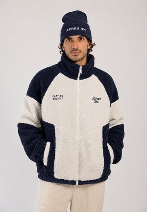 Homme portant une veste polaire bleu marine et blanche avec fermeture éclair à l'avant et un bonnet bleu marine sur lequel il est écrit "APRÈS SKI" sur fond gris uni.