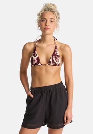 Bikini met een kastanjebruine basis en crèmekleurige bloemenpatronen, met halterbandjes die strikken en een houten accent; gecombineerd met gestructureerde zwarte shorts.