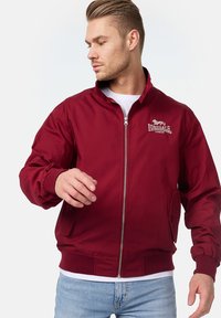 Bordo bomber jakna sa zatvaračem sprijeda, rebrastim manžetama i ovratnikom. Sadrži izvezeni logo na prsima i dva bočna džepa.