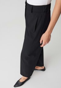 Pantalons noirs taille haute avec une coupe décontractée, dotés de poches latérales et de plis cousus. Associés à des chaussures noires à bout pointu.