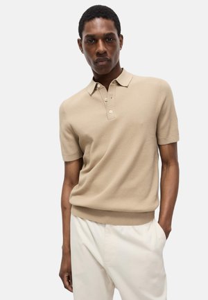 Polo shirt beige a maniche corte con un motivo testurizzato, colletto con tre bottoni e orlo a coste. Indossato con pantaloni di colore chiaro.