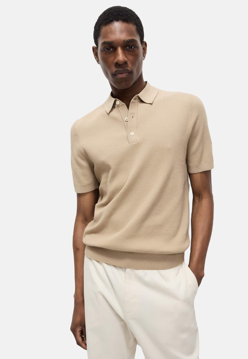 Polo de manga corta beige con un patrón texturizado, tapeta de tres botones y dobladillo acanalado. Se lleva con pantalones de color claro.