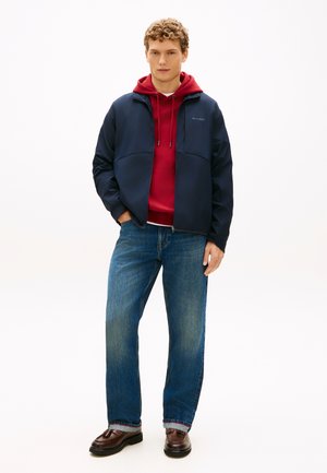 Marineblauer Windbreaker mit Kapuze über rotem Kapuzenpullover, kombiniert mit blauen Jeans und braunen Loafers. Verfügt über Reißverschluss und Logo auf der Brust.