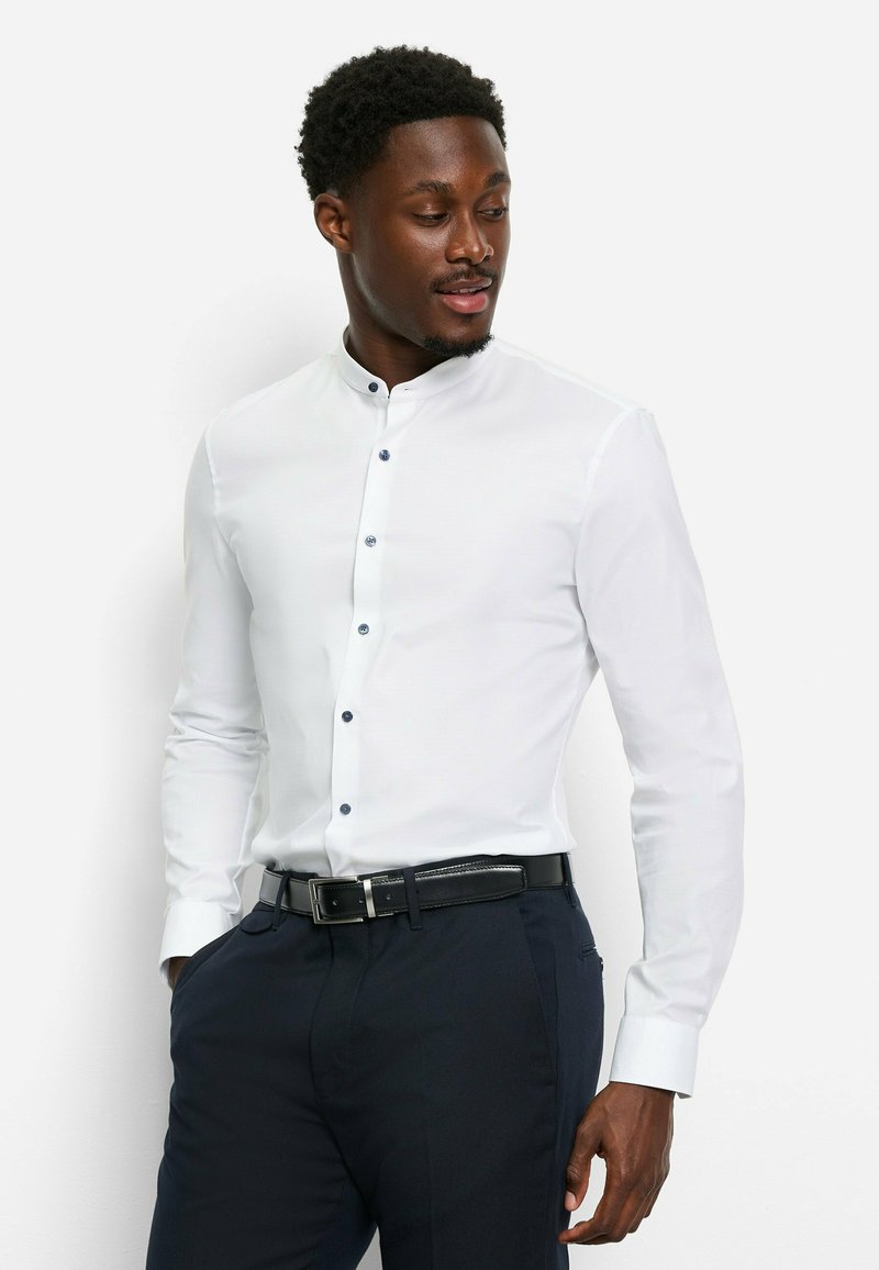 Weißes Hemd mit langen Ärmeln, Stehkragen und blauen Knöpfen, kombiniert mit dunklen Hosen und einem schwarzen Gürtel, in einer taillierten Schnittführung.