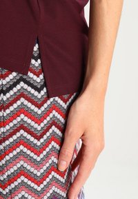 Primer plano de una mano reposando sobre una falda de patrón zigzag multicolor, con una blusa burdeos que presenta una abertura lateral.