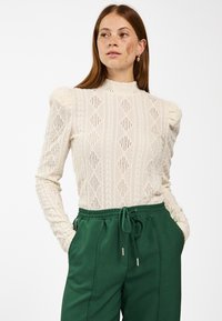 Gehaakte crèmekleurige lange mouw top met ruitpatronen en textuurdetails, gecombineerd met groene broek met tailleband in een ontspannen pasvorm.