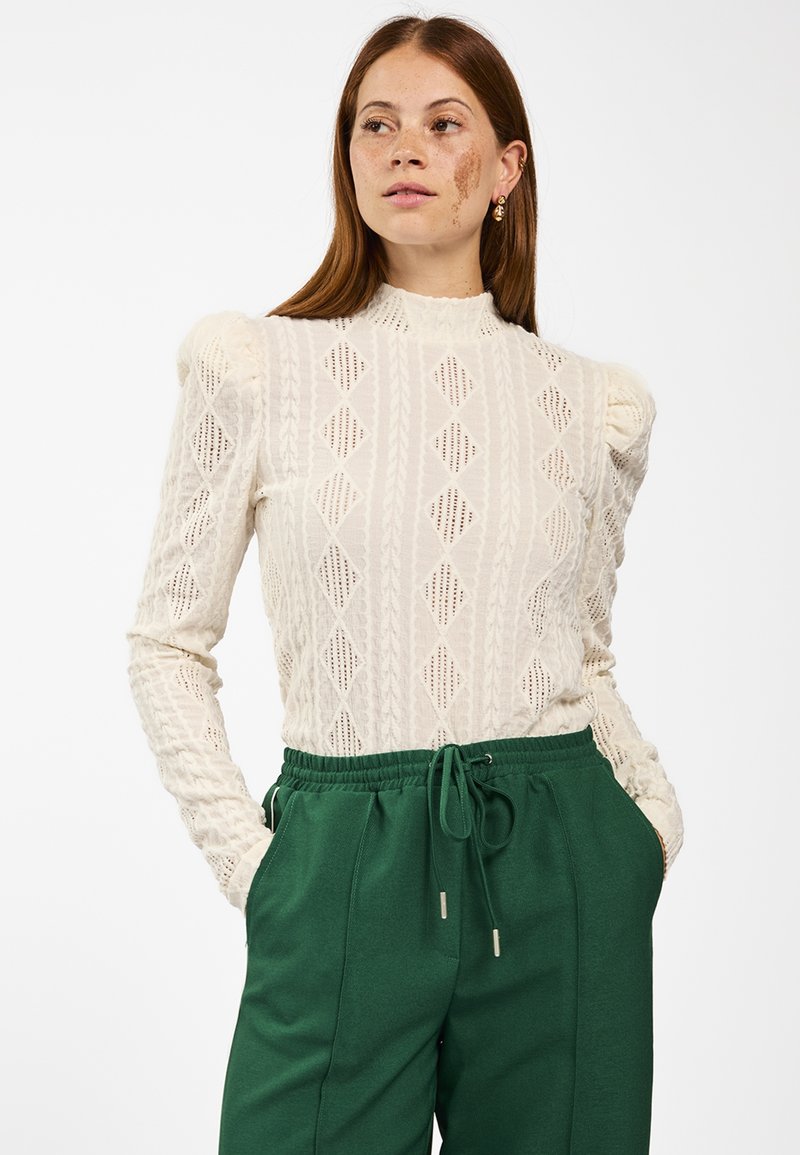 Gehaakte crèmekleurige lange mouw top met ruitpatronen en textuurdetails, gecombineerd met groene broek met tailleband in een ontspannen pasvorm.