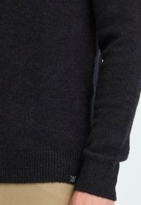 Donkergrijze trui van zacht materiaal, met een geribde rand en mouwen. Klein logo detail aan de onderkant aan de linkerkant.