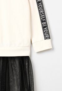 Sudadera crema con texto negro en la manga, combinada con una falda de tul negro semitransparente, que presenta una textura suave y un corte relajado.