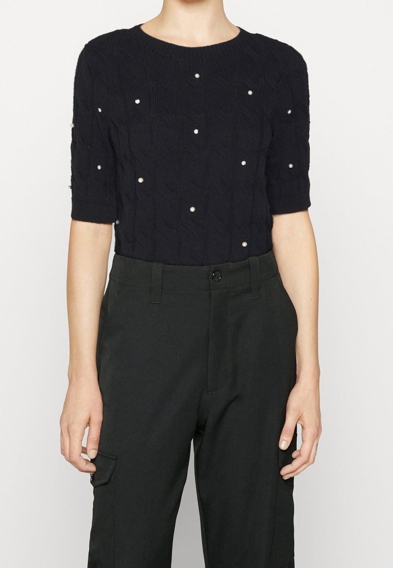 Haut en maille noir à manches courtes avec des ornements en perles, associé à un pantalon noir taille haute avec poches latérales, porté par une personne debout.