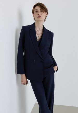 Persona con capelli corti che indossa un abito blu navy su misura, in piedi contro un muro bianco con una mano in tasca e un'espressione neutra.
